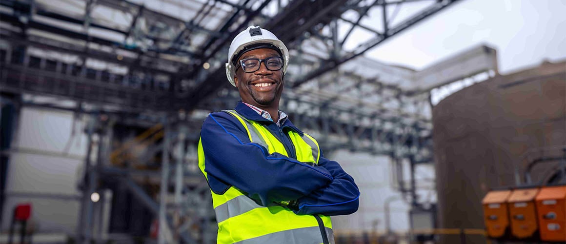 A smiling man wearing a white hard hat, glasses, and a yellow reflective vest is standing with crossed arms at Elikhulu mine.